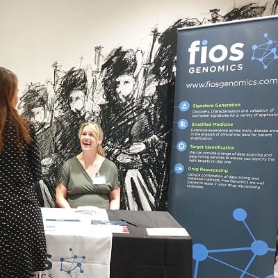 Fios Genomics Stand at Genomic Medicine Edinburgh 2019. Eleanore is sat behiind the table talking to a woman about our services.