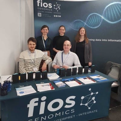 5 members of the Fios team at the Festival of Genomics 2020. The Fios backdrop is behind them, 3 of the team are standing up and 2 are sitting down. The table has Fios-branded notepads, pens and flyers on it.